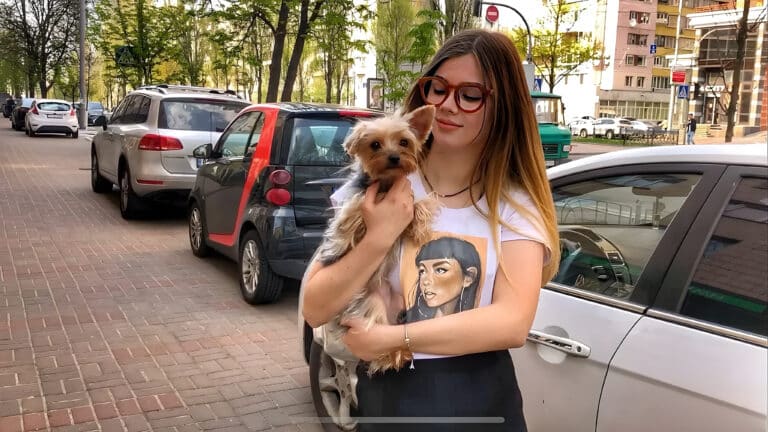 Дівчина в окулярах стоїть біля срібної машини на вулиці, тримаючи в руках йорка