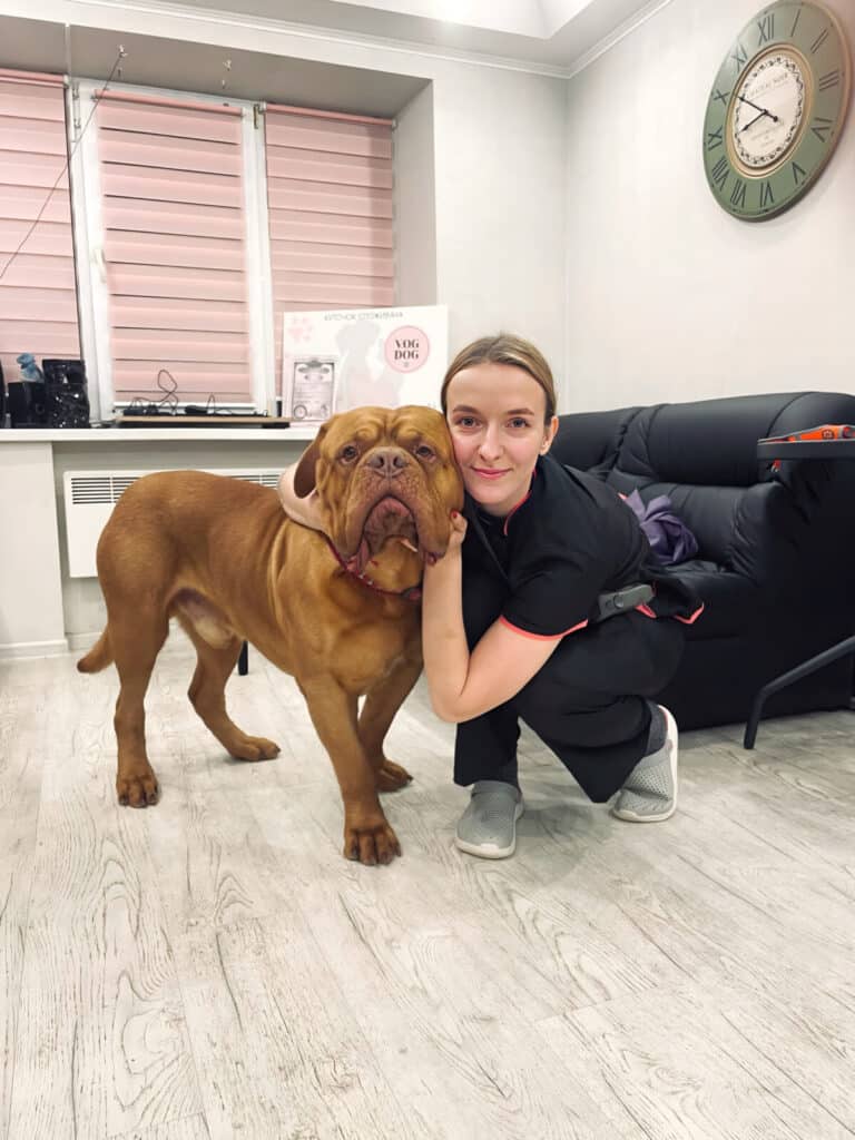 Грумерка в сірих капцях обійняла на підлозі коричневого великого пса біля чорного дивана