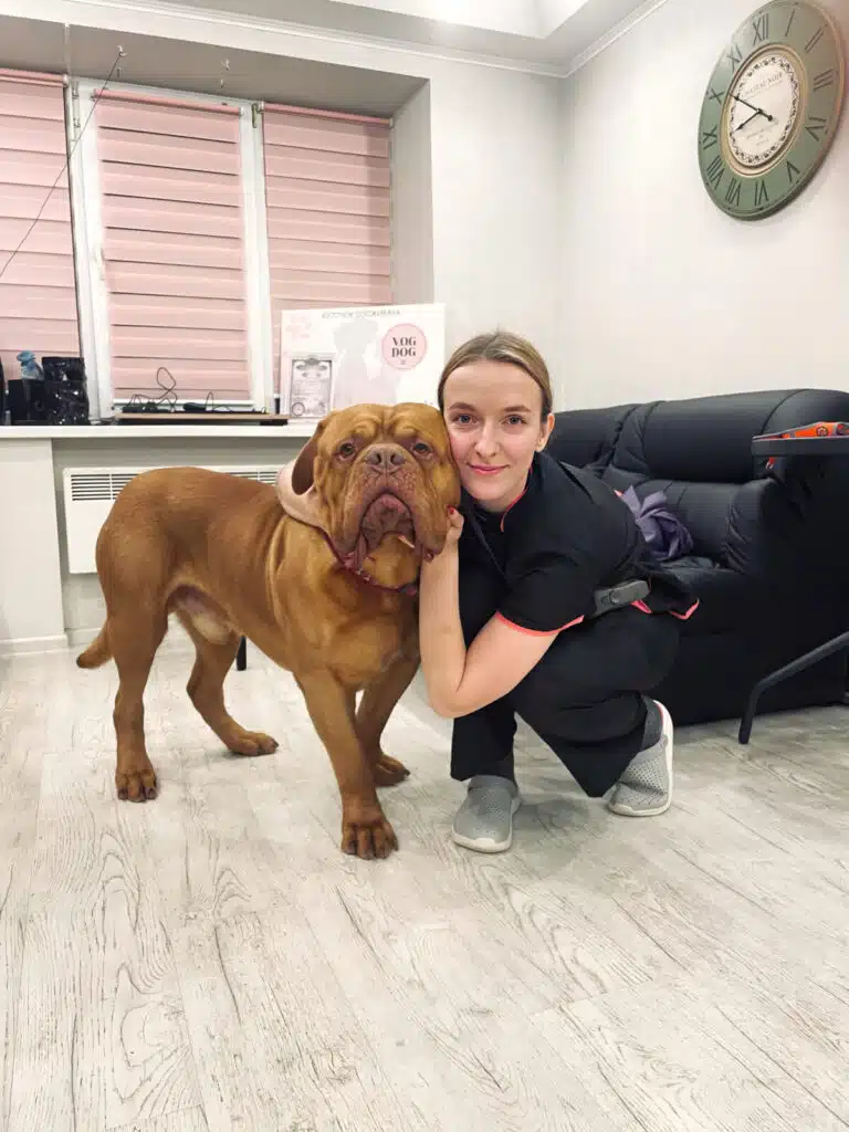 Грумерка в сірих капцях обійняла на підлозі коричневого великого пса біля чорного дивана