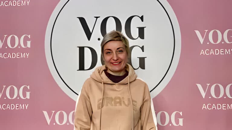 Жінка з коротким волоссям стоїть перед великою вивіскою Академії, вдягнута в світло-коричневе худі