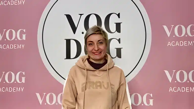 Жінка з коротким волоссям стоїть перед великою вивіскою Академії, вдягнута в світло-коричневе худі