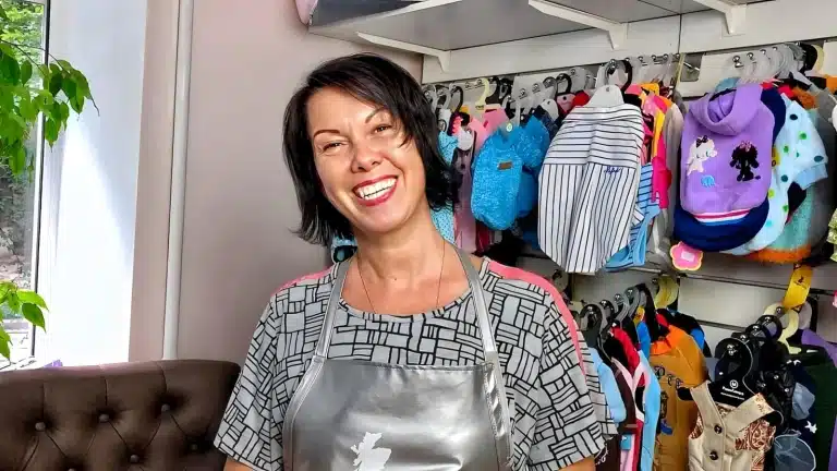 Жінка з чорним каре сміється на камеру біля стенда з одягом