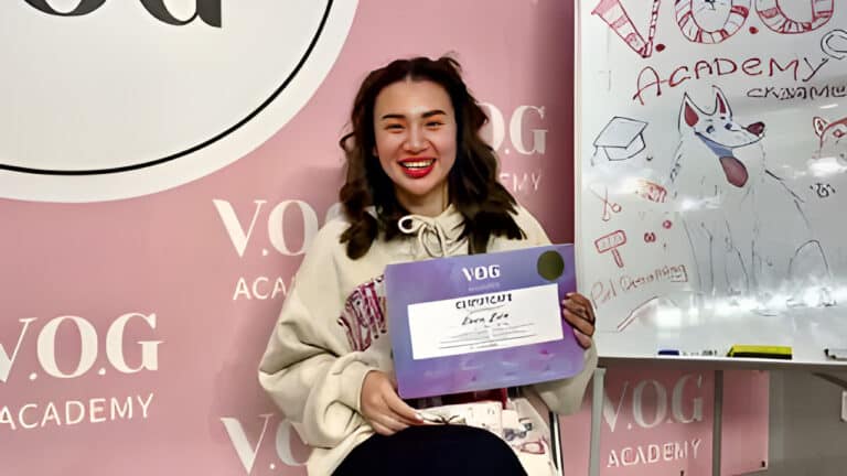 Дівчина з вузькими очима і червоними губами сидить біля дошки, тримаючи фіолетовий сертифікат