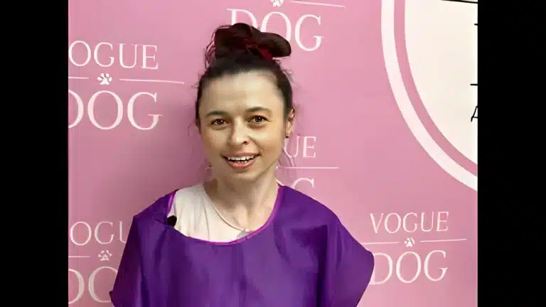 Жінка в бежевій футболці та фіолетовому фартусі стала біля рожевої стіни з написами та привідкрила рота