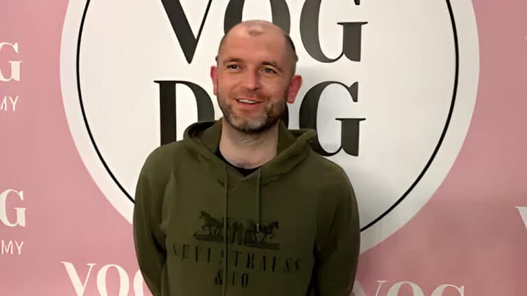 Чоловік в темно-зеленому худі склав за спиною руки та дивиться в бік