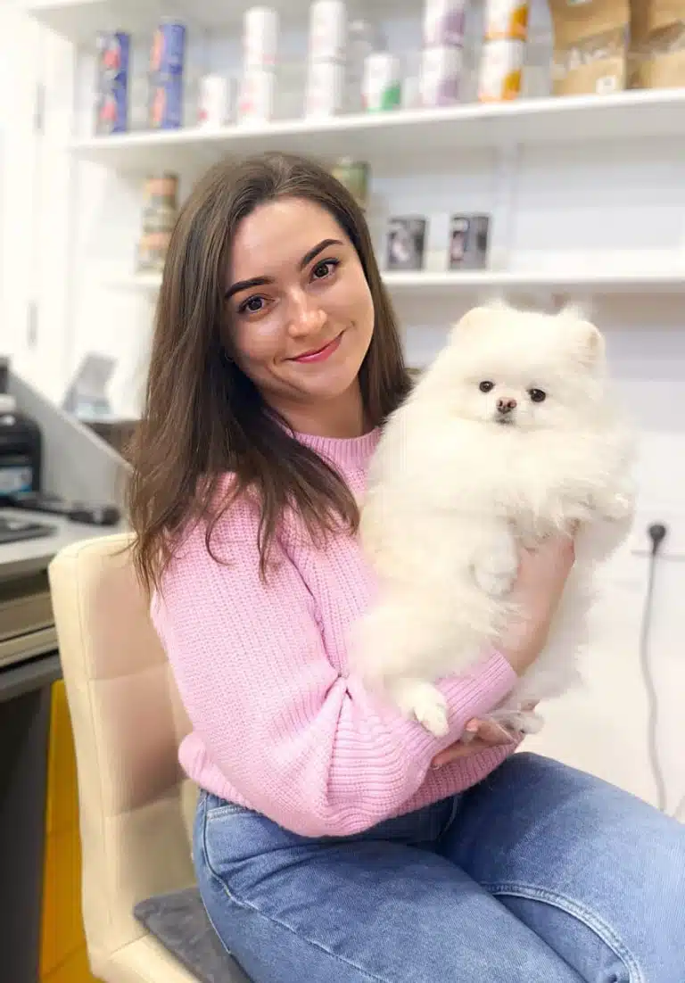 Дівчина в рожевому светрі однією рукою тримає під грудку білого шпіца з густою шерстю