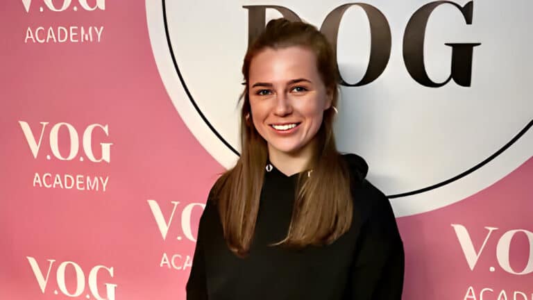 Рудоволоса дівчина з круглими сережками всміхається на камеру на фоні рожевого банера Академії