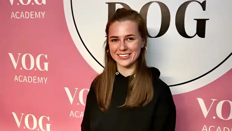 Рудоволоса дівчина з круглими сережками всміхається на камеру на фоні рожевого банера Академії