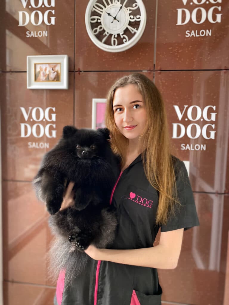Дівчина у формі грумінг-салону стоїть перед білим годинником, тримаючи в руках чорного шпіца