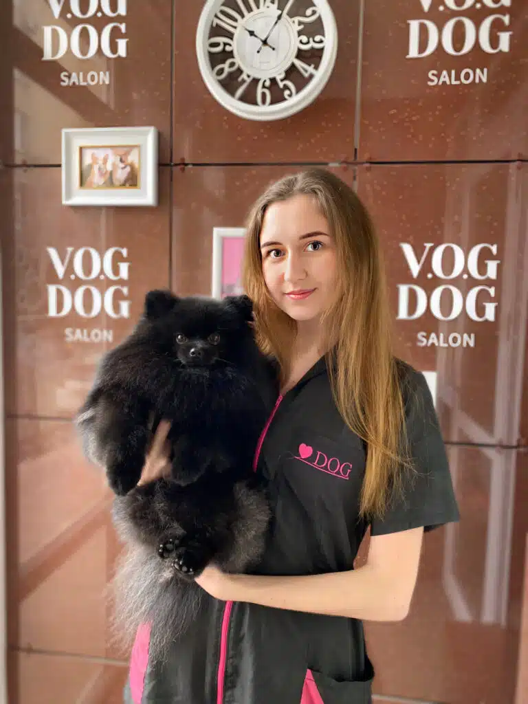 Дівчина у формі грумінг-салону стоїть перед білим годинником, тримаючи в руках чорного шпіца