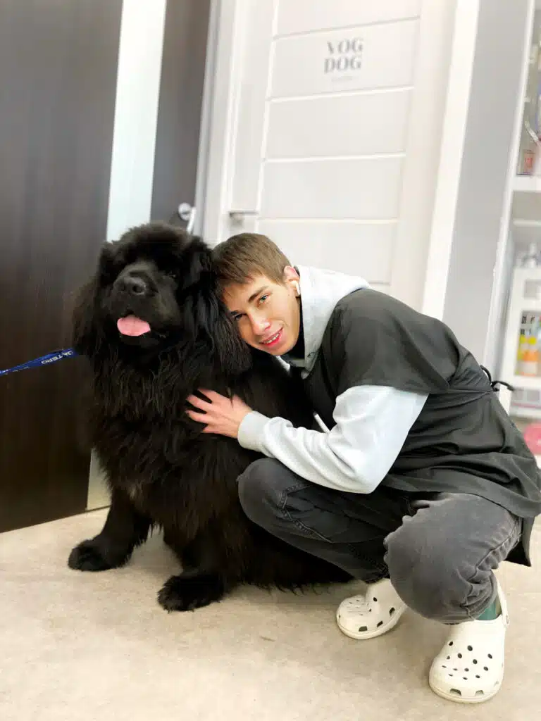 Хлопець в білих гумових капцях обіймає на підлозі великого пухнастого пса чорного забарвлення