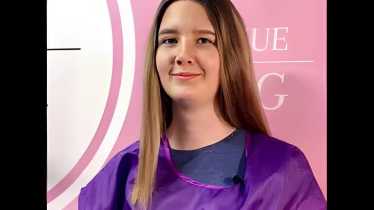 Дівчина з рівним волоссям та у фіолетовому фартусі посміхається на камеру перед рожевим банером