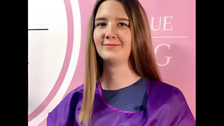 Дівчина з рівним волоссям та у фіолетовому фартусі посміхається на камеру перед рожевим банером