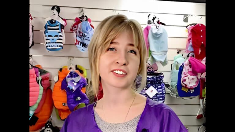 Дівчина зі світлим волоссям одягла на себе фіолетову накидку та стала перед стіною з одягом на продаж