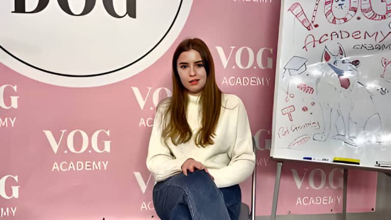 Рудоволоса дівчина в білому светрі сидить біля дошки з малюнком