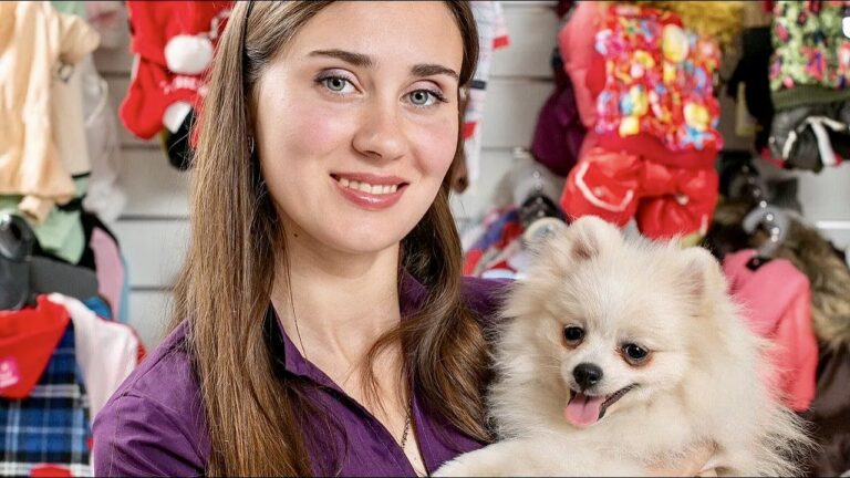 Дівчина у фіолетовій формі тримає на руках кремового шпіца з коричневою шерстю під очима на фоні стенда з одягом