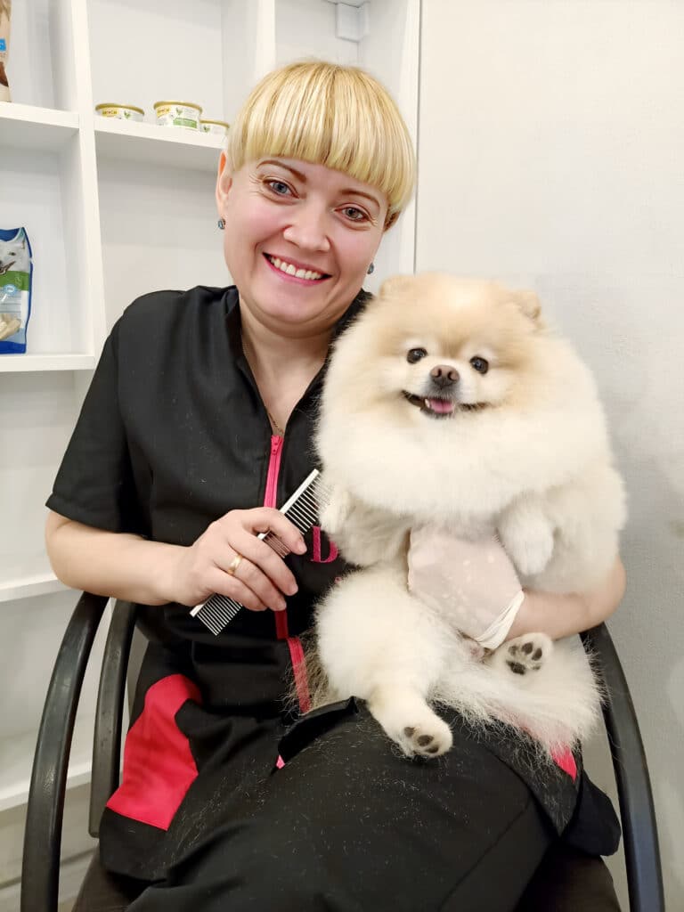 Білий шпіц та грумер з білою гривкою посміхаються разом біля стенда зі смаколиками