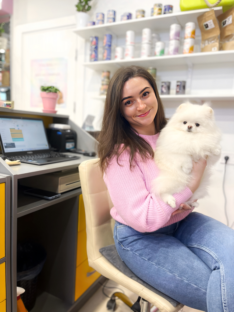 Дівчина в рожевому светрі сидить спиною до ноутбука, тримаючи в руках кремового шпіца
