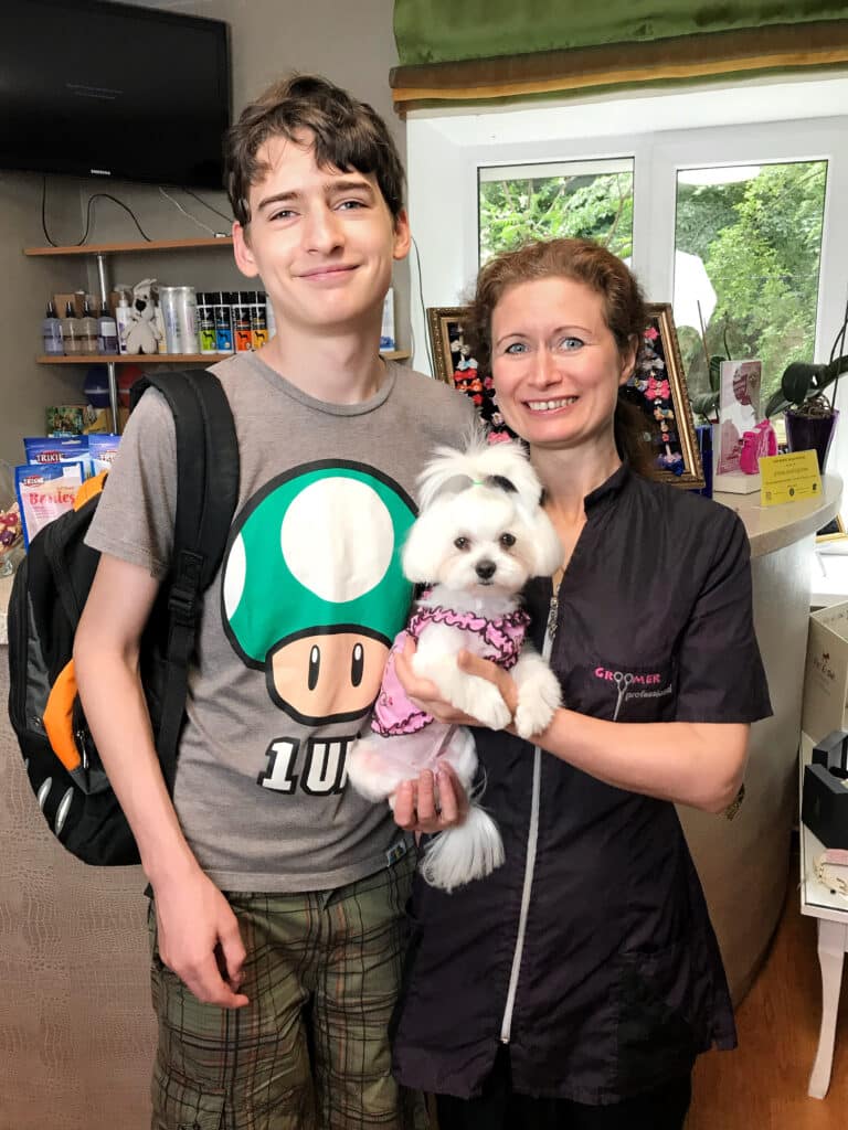 Грумер у формі тримає в руках мальтезе в рожевій сукні, стоячи поруч з високим хлопцем