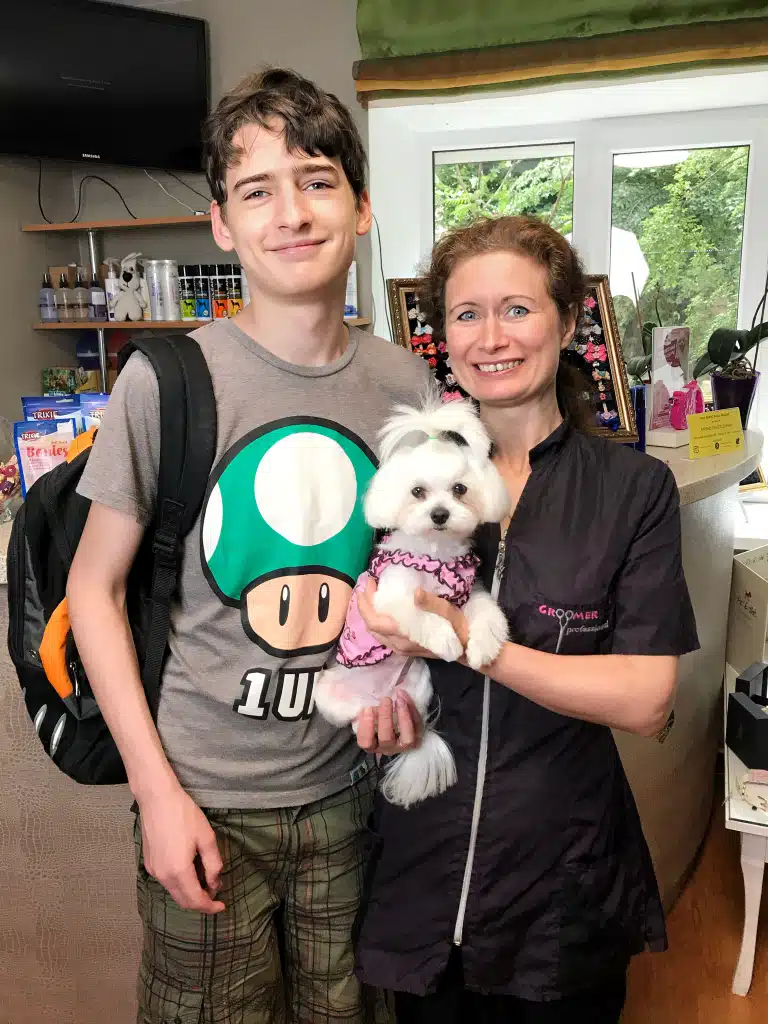 Грумер у формі тримає в руках мальтезе в рожевій сукні, стоячи поруч з високим хлопцем