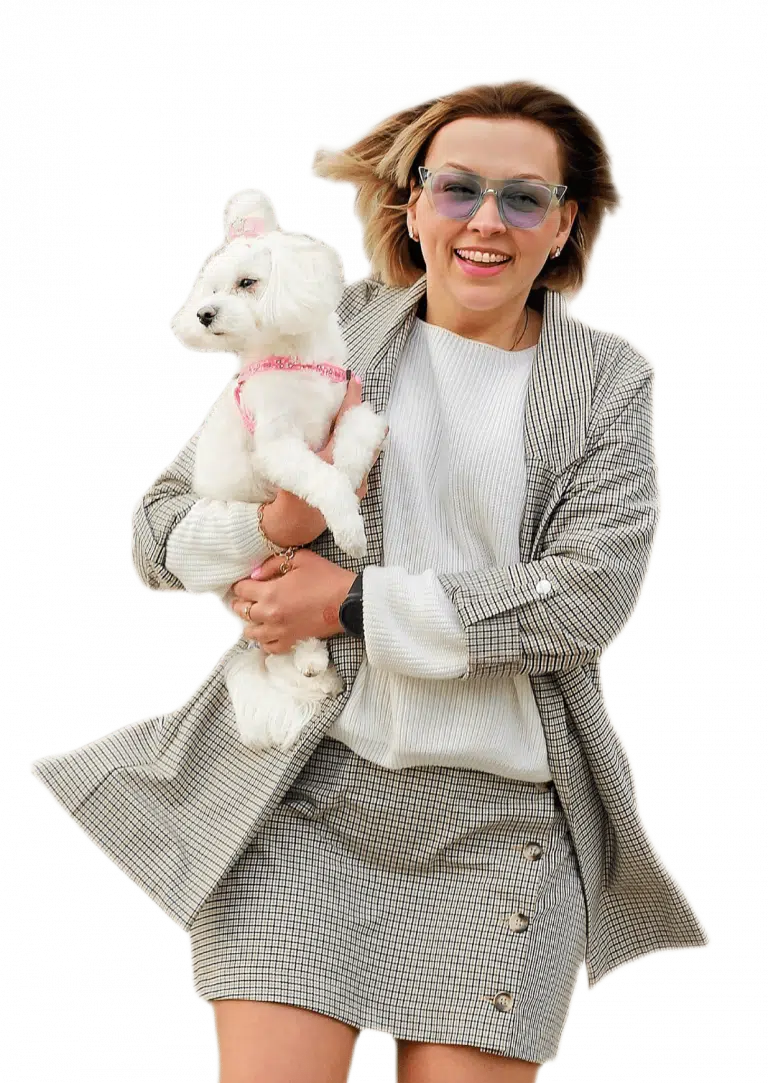 Дівчина в прозорих окулярах з блакитним склом посміхається, тримаючи мальтезе в рожевій амуніції на руках