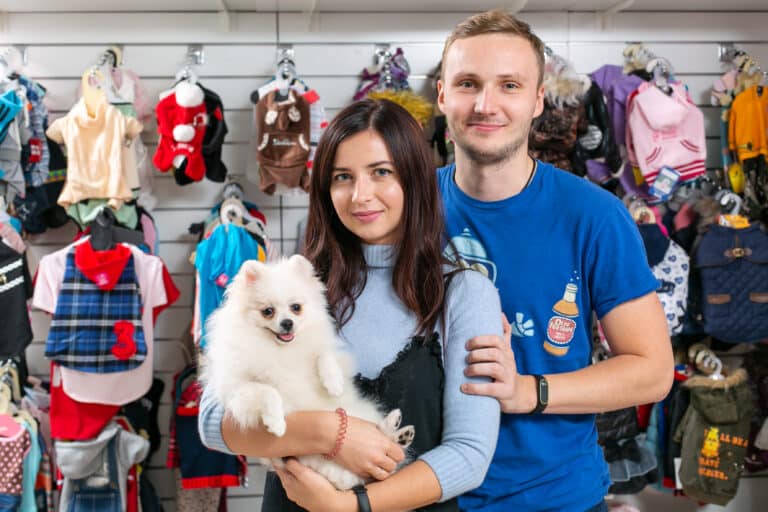 Білий шпіц сидить на руках дівчини в блакитному светрі та чорній сукні, які приобіймає молодий чоловік