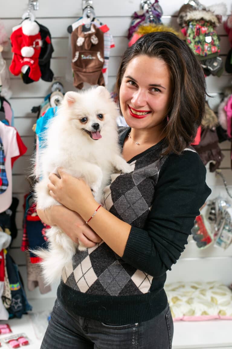Дівчина з червоною помадою тримає в руках білого шпіца на фоні стенда з різним одягом для собак