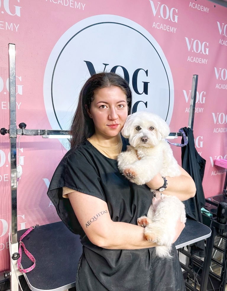 Дівчина з татуюванням вище ліктя тримає на руках кремового підстриженого мальтіпу на фоні рожевого банера