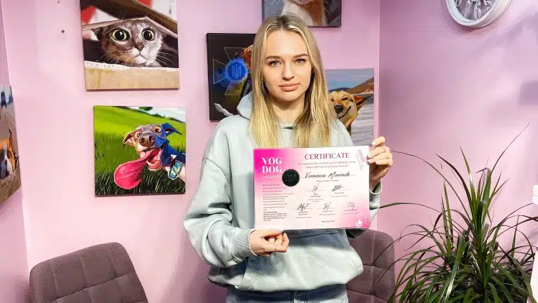 Дівчина зі світлим волоссям тримає в руках рожевий сертифікат Академії грумінгу біля зеленого вазона