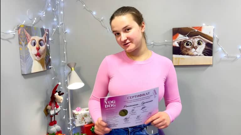 Дівчина в рожевому гольфі тримає сертифікат закінчення курсів Академії грумінгу