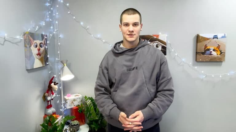 Посеред сірої кімнати стоїть високий хлопець в сірому худі з чорним написом на грудях