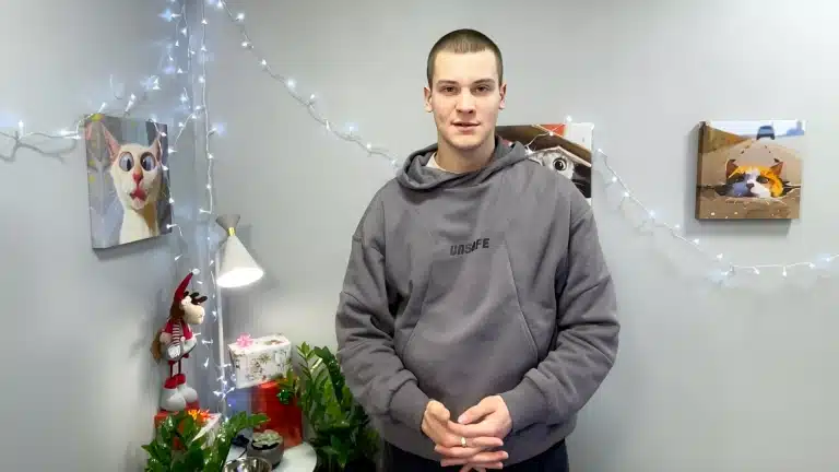 Посеред сірої кімнати стоїть високий хлопець в сірому худі з чорним написом на грудях