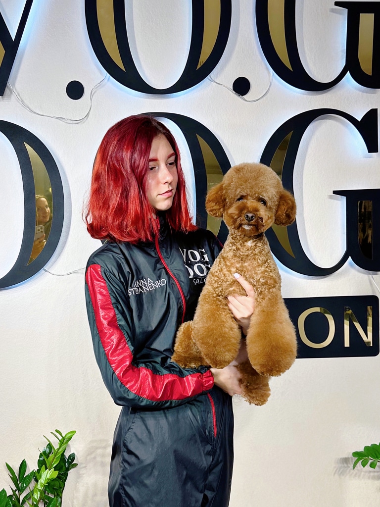 Дівчина з червоним волоссям тримає в руках мальтіпу з об'ємними ніжками біля світлої стіни