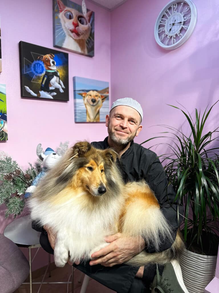 Чоловік в грумерській формі тримає на руках пухнастого коллі, сидячи біля зеленого вазона