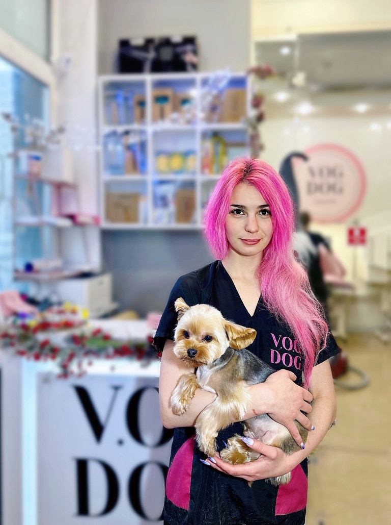Грумер з довгим рожевим волоссям тримає на руках підстриженого йорка перед входом в грумерську