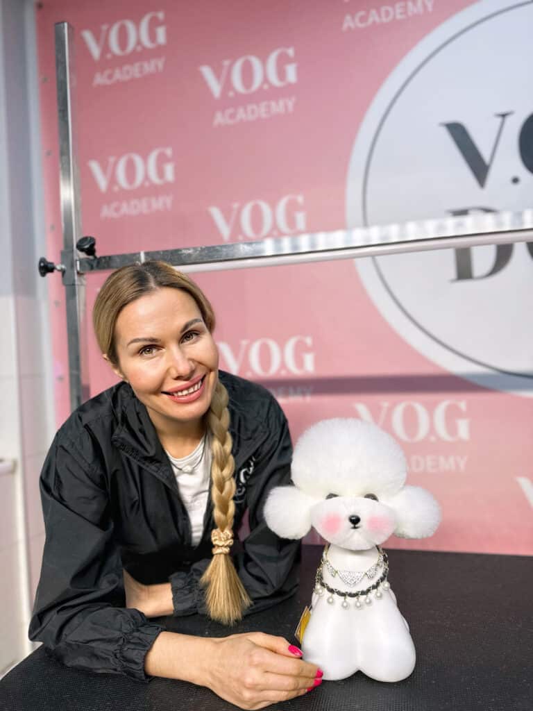 Жінка з довгою косою сперлась на чорний стіл, на якому стоїть підстрижений моделдог