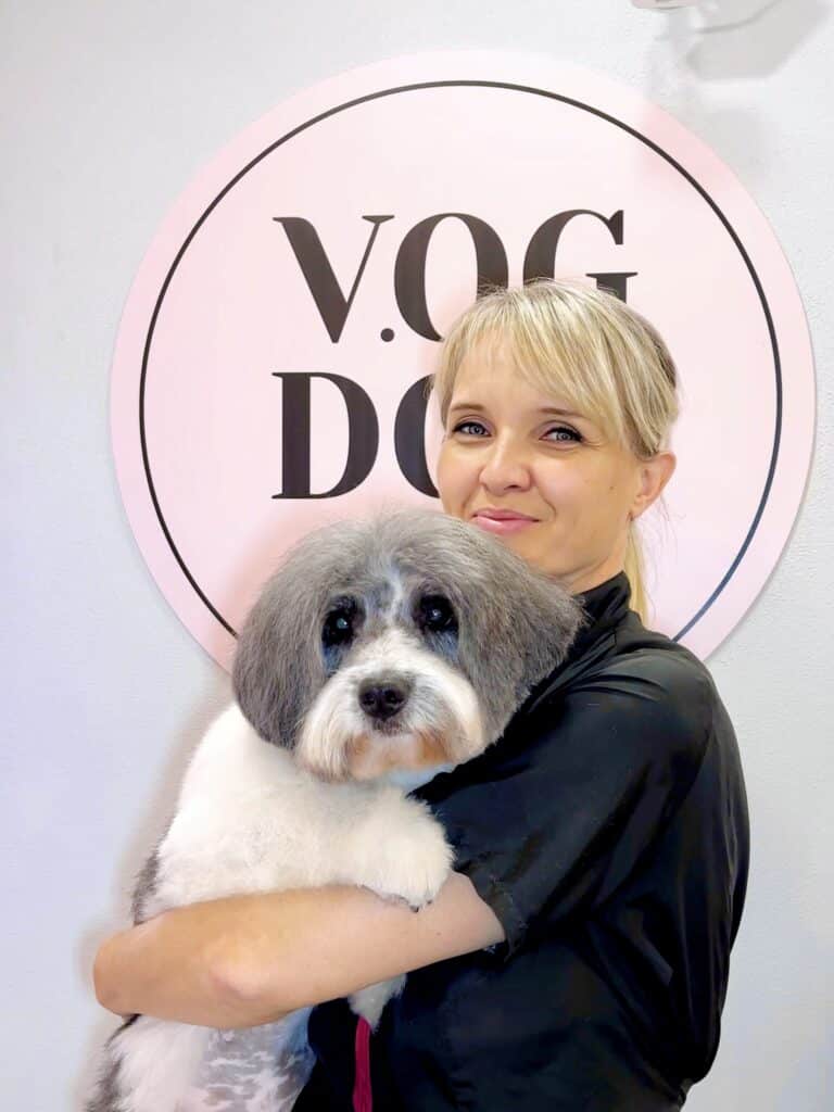 Грумерка зі світлим волоссям тримає великого та пухнастого пса перед рожевим банером салону