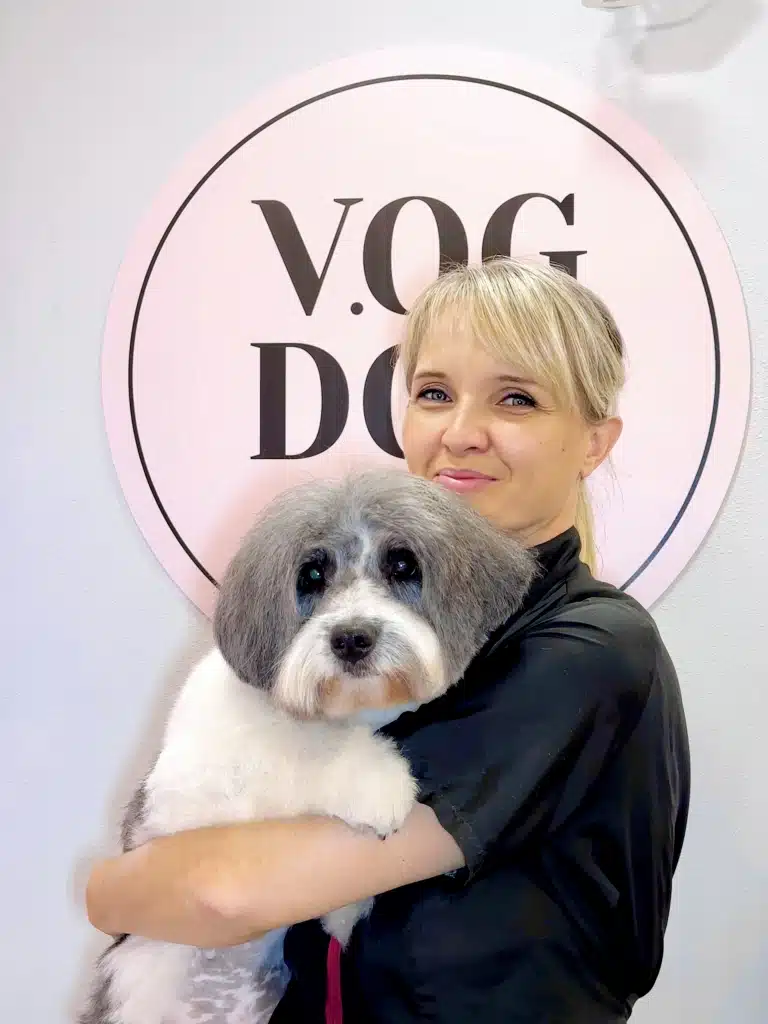 Грумерка зі світлим волоссям тримає великого та пухнастого пса перед рожевим банером салону