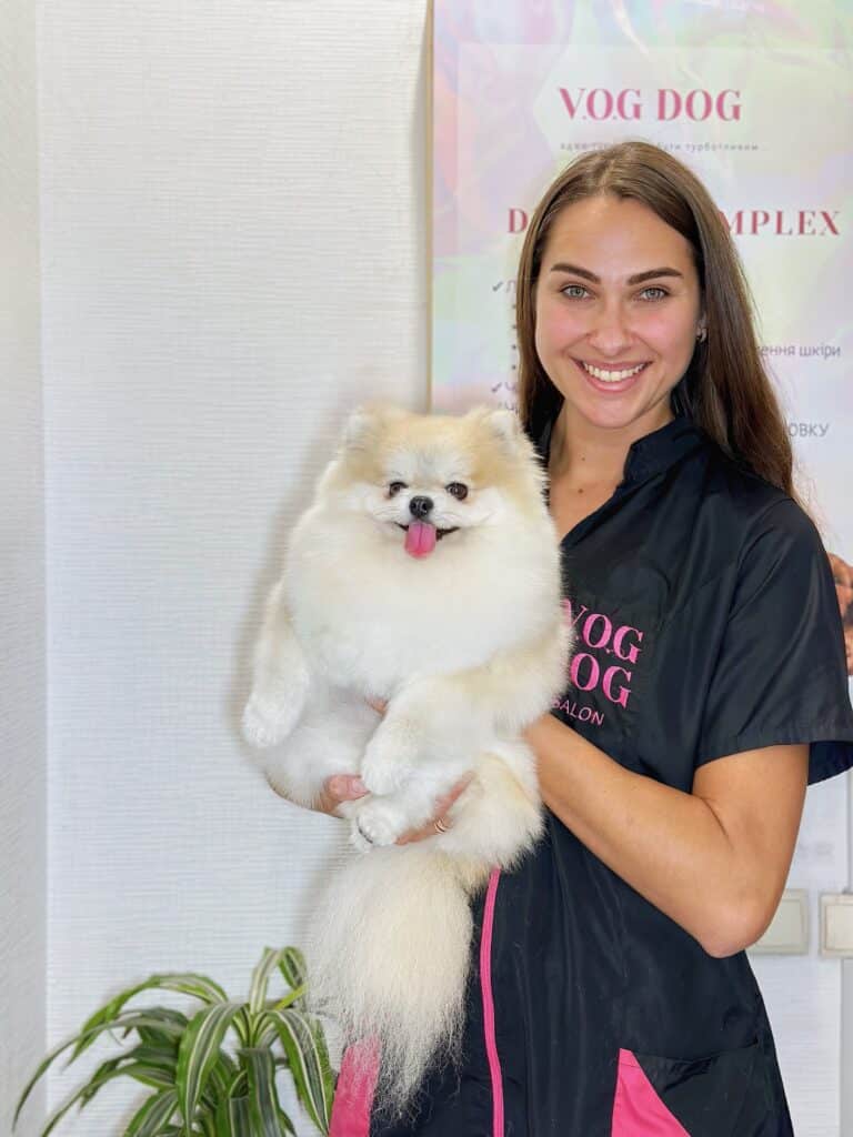 Грумер з рівним волоссям посміхається разом зі шпіцом на руках у світлій кімнаті