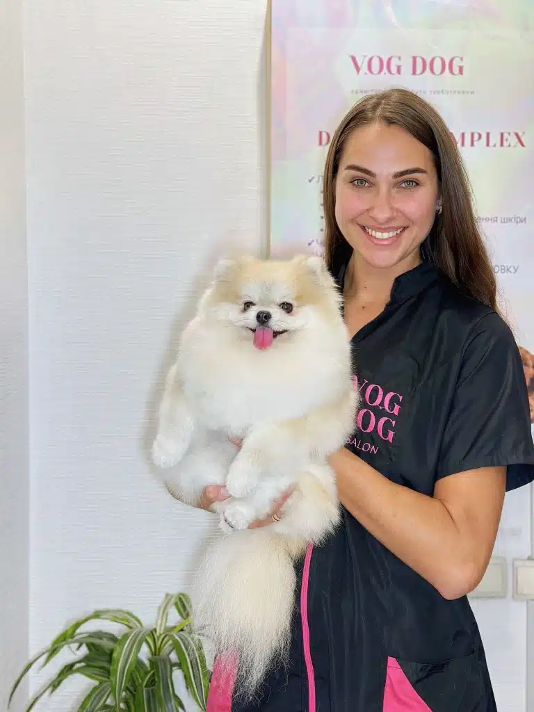 Грумер з рівним волоссям посміхається разом зі шпіцом на руках у світлій кімнаті