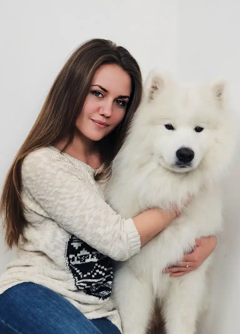 Дівчина з коричневим волоссям обіймає обома руками білого самоїда