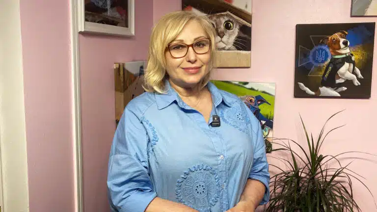 Жінка в блакитній сорочці має коротке світле волосся та гарні окуляри