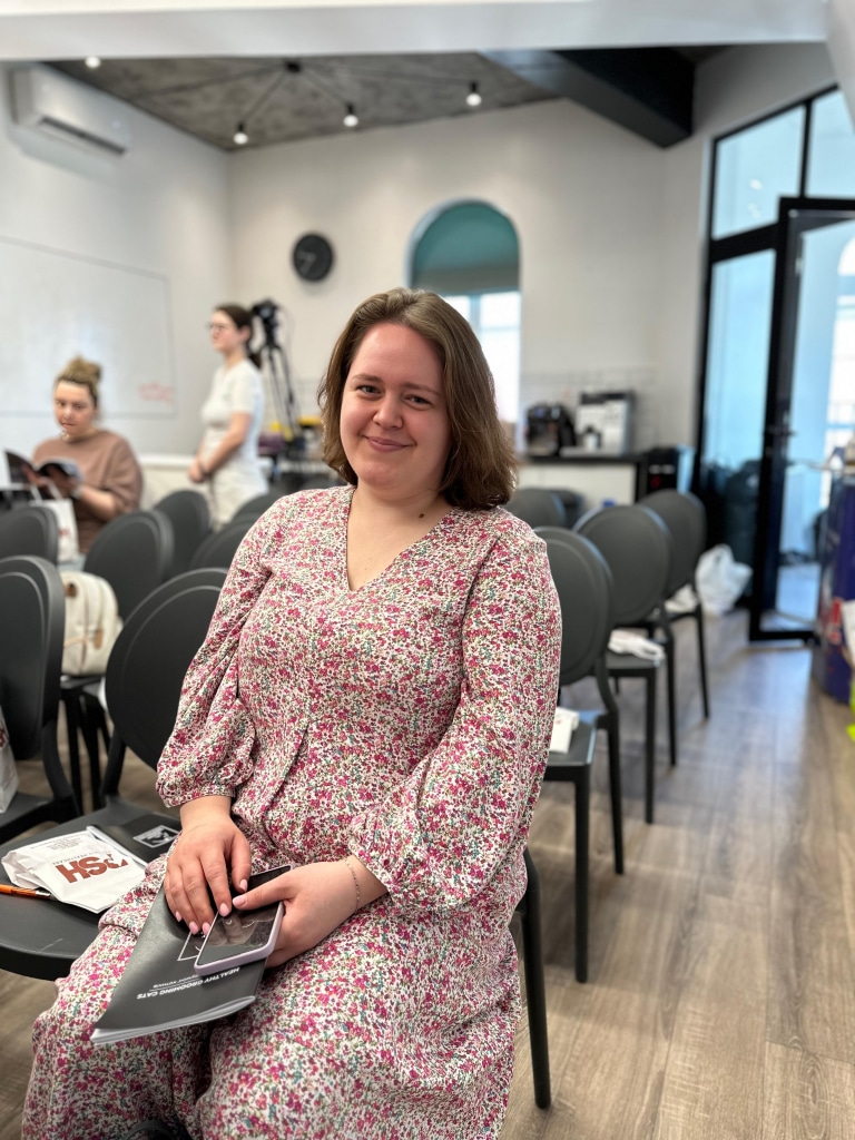 Дівчина з короткою стрижкою у сукні з квітами тримає в руках нотатник та телефон