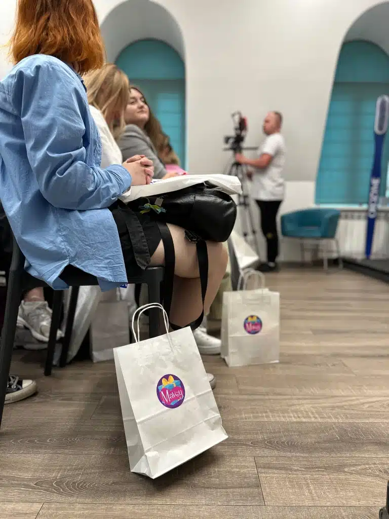 Поруч з дівчатами, що зайняли перший ряд, лежать білі крафтові пакети з логотипом бренду