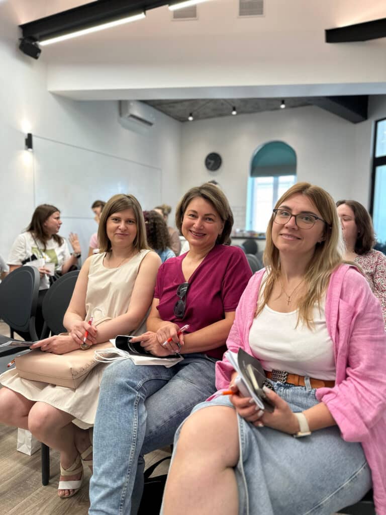 Троє жінок в одежі біло-рожевого кольору сидять разом та посміхаються