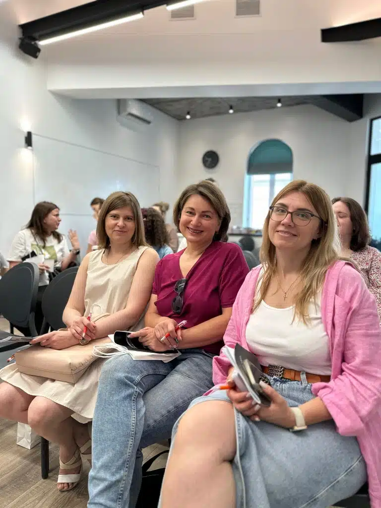 Троє жінок в одежі біло-рожевого кольору сидять разом та посміхаються