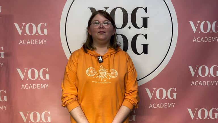 Жінка в помаранчевій кофтинці з капюшоном стоїть перед рожевим банером