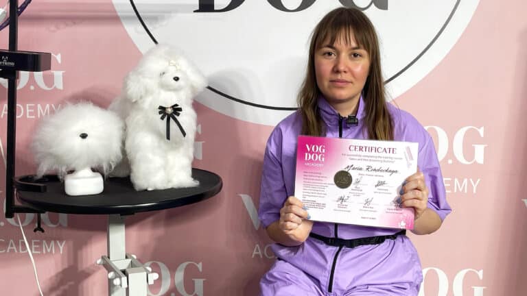 Дівчина в фіолетовій формі тримає сертифікат в руках, сидячи біля моделей для стрижки