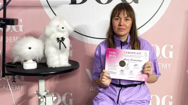 Дівчина в фіолетовій формі тримає сертифікат в руках, сидячи біля моделей для стрижки