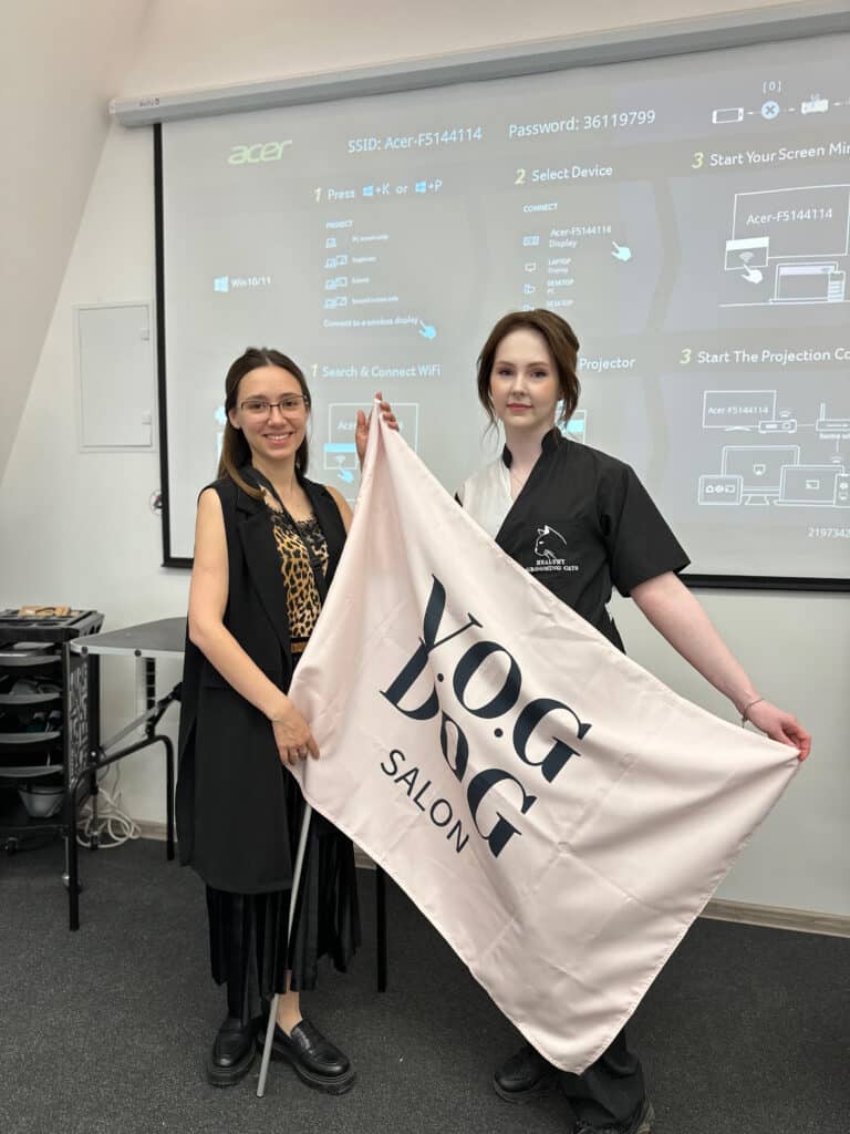 Дівчина в довгій чорній жилетці тримає стяг салону грумінгу разом з викладачем лекції